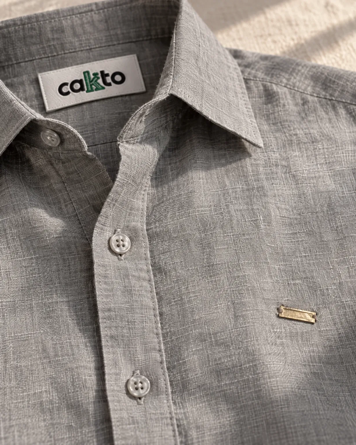 Detalle de la tela, la plaquita metálica y la etiqueta Cakto de la camisa 100% lino gris