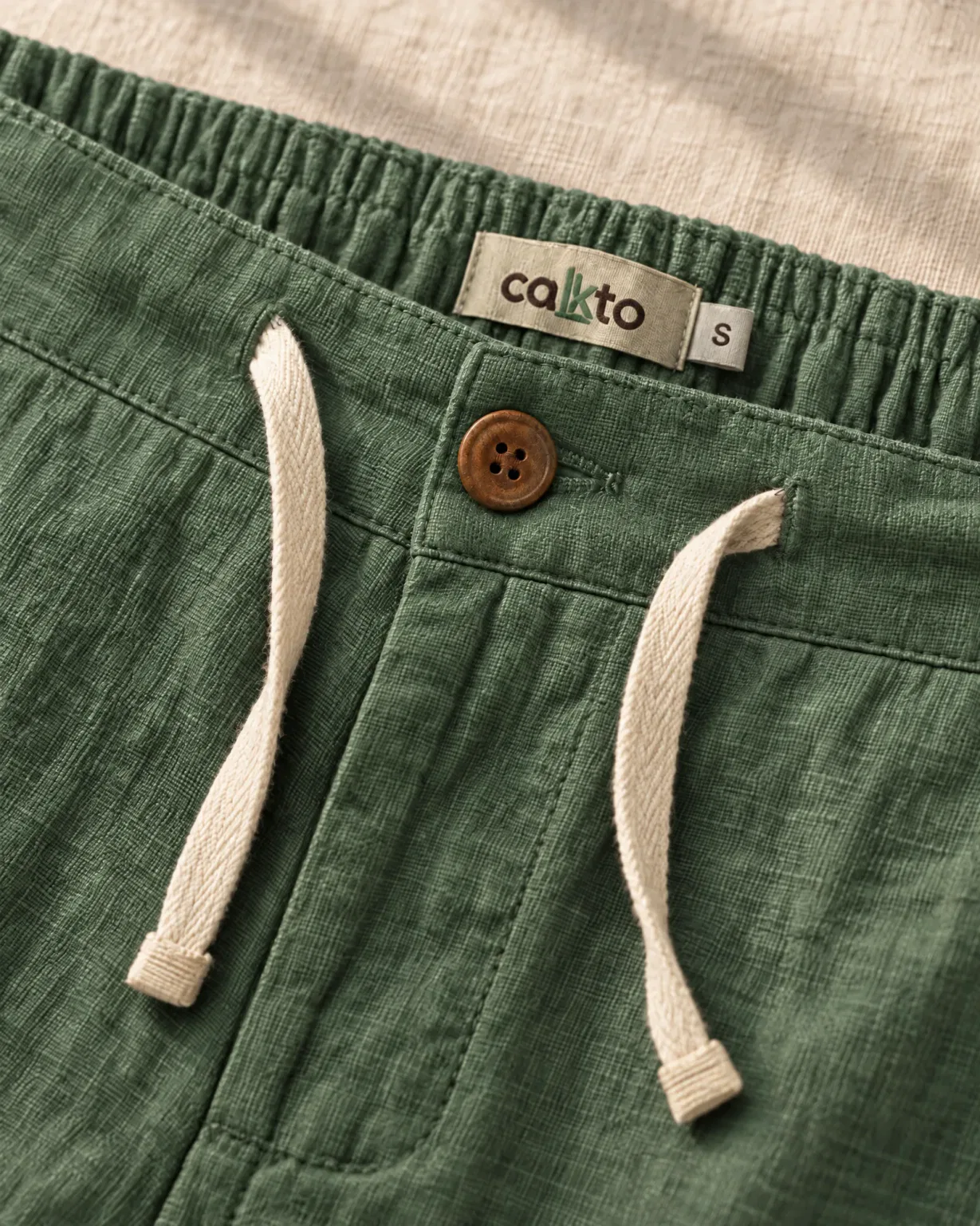 Detalle de la pretina con botón, cordón y marquilla Cakto del pantalón de lino verde